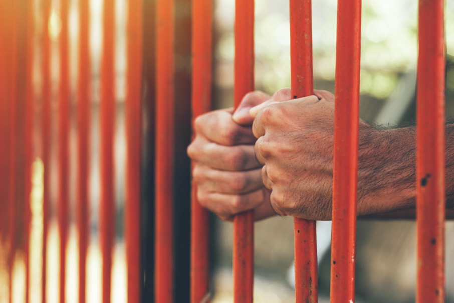 A imagem mostra mãos atrás das grades de uma prisão. A imagem ilustra o texto "Auxílio Reclusão: regras e valores para pagamento do benefício" de Koetz Advocacia.