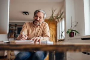 A imagem mostra um homem de meia-idade, sentado à mesa, escrevendo em um caderno. Ilustra o texto sobre Planejamento de aposentadoria.