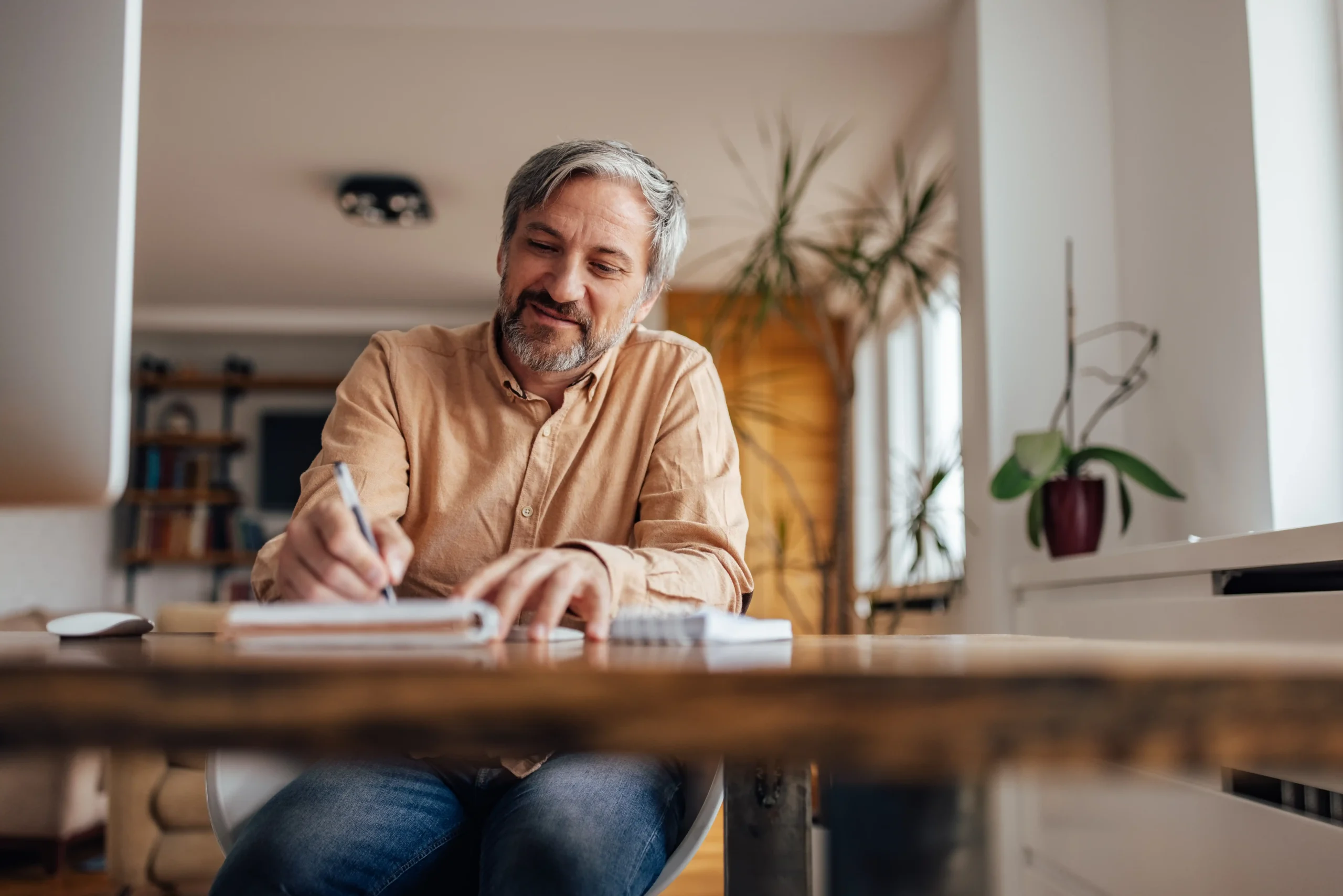 A imagem mostra um homem de meia-idade, sentado à mesa, escrevendo em um caderno. Ilustra o texto sobre Planejamento de aposentadoria.