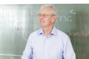 A imagem mostra um professor sênior, dentro da sala de aula em frente à lousa. Ilustra o texto sobre aposentadoria do professor.