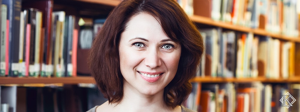 Jovem professora sorrindo em frente à estante com diversos livros. A imagem foi escolhida para representar a importância de se antecipar e realizar o planejamento de aposentadoria de professor.