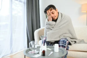 A imagem mostra um jovem homem doente, enrolado em um cobertor, enquanto está sentado em um sofá.