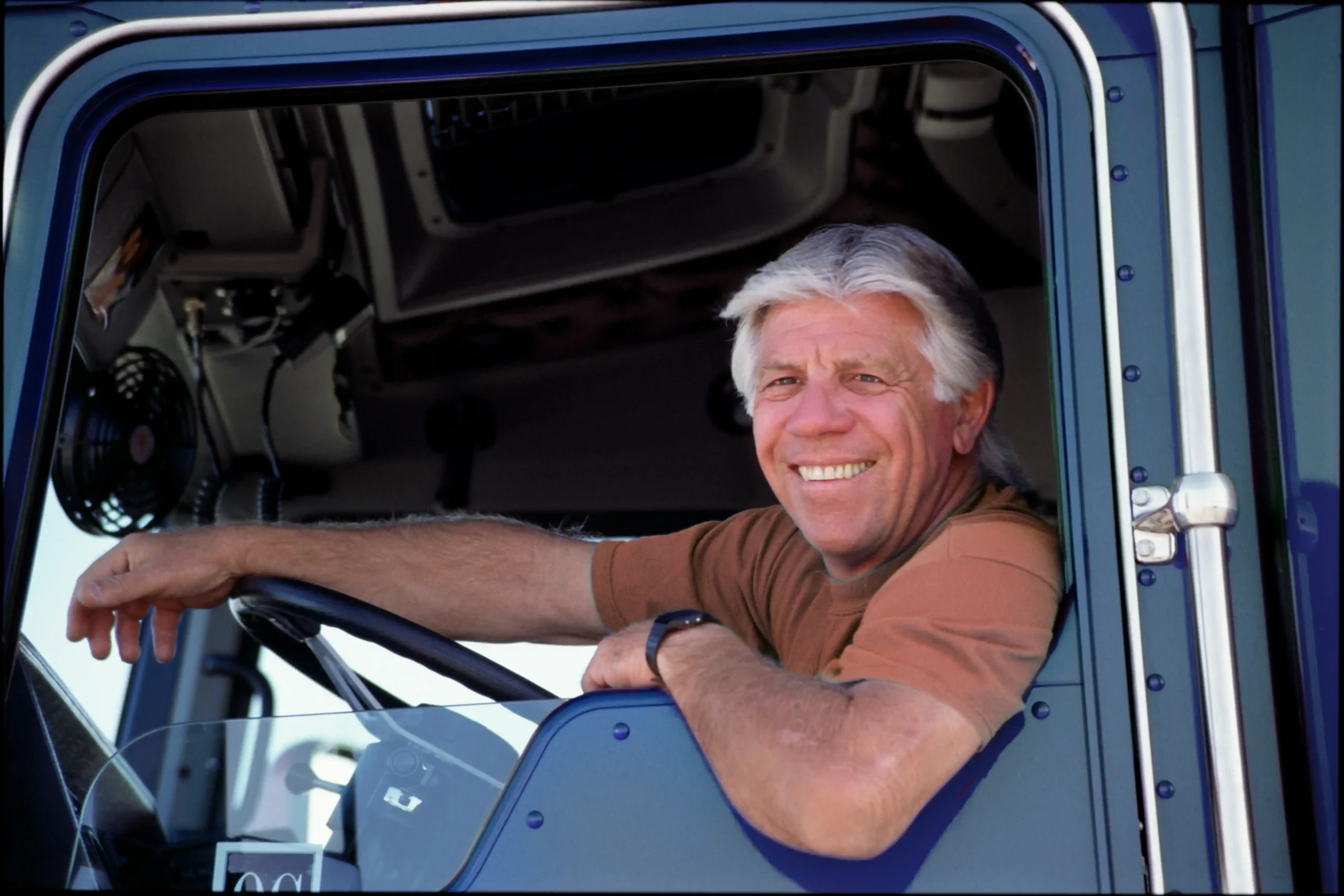 A imagem mostra um motorista de caminhão, sentado em frente ao volante, apoiando um dos braços na janela aberta e sorrindo para a câmera. A imagem ilustra a aposentadoria do caminhoneiro.