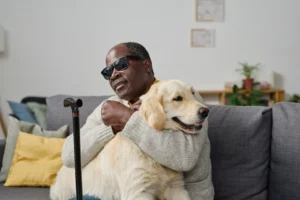 A imagem mostra um homem cego, de meia idade, usando óculos escuros, sentado em um sofá, enquanto abraça seu cão guia. Ilustra o texto se cegueira aposenta.