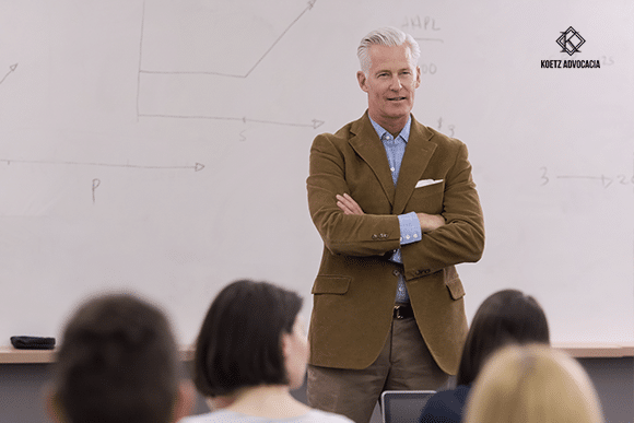 A imagem mostra um professor dando sua aula em uma sala.