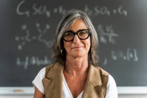 A imagem mostra uma professora de meia-idade, em frente à lousa, sorrindo para a câmera. Ilustra o texto sobre Auxílio doença para professores.