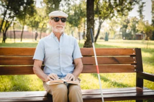 A imagem mostra um homem sênior, cego, ao ar livre, sentado em um banco, lendo um livro em braile, enquanto sorri. Ilustra o texto sobre Aposentadoria da pessoa com deficiência.