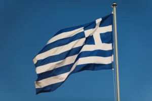 A imagem mostra a bandeira da Grécia hasteada, balançando com o vento. Ilustra o texto sobre Acordo Previdenciário Internacional Grécia.