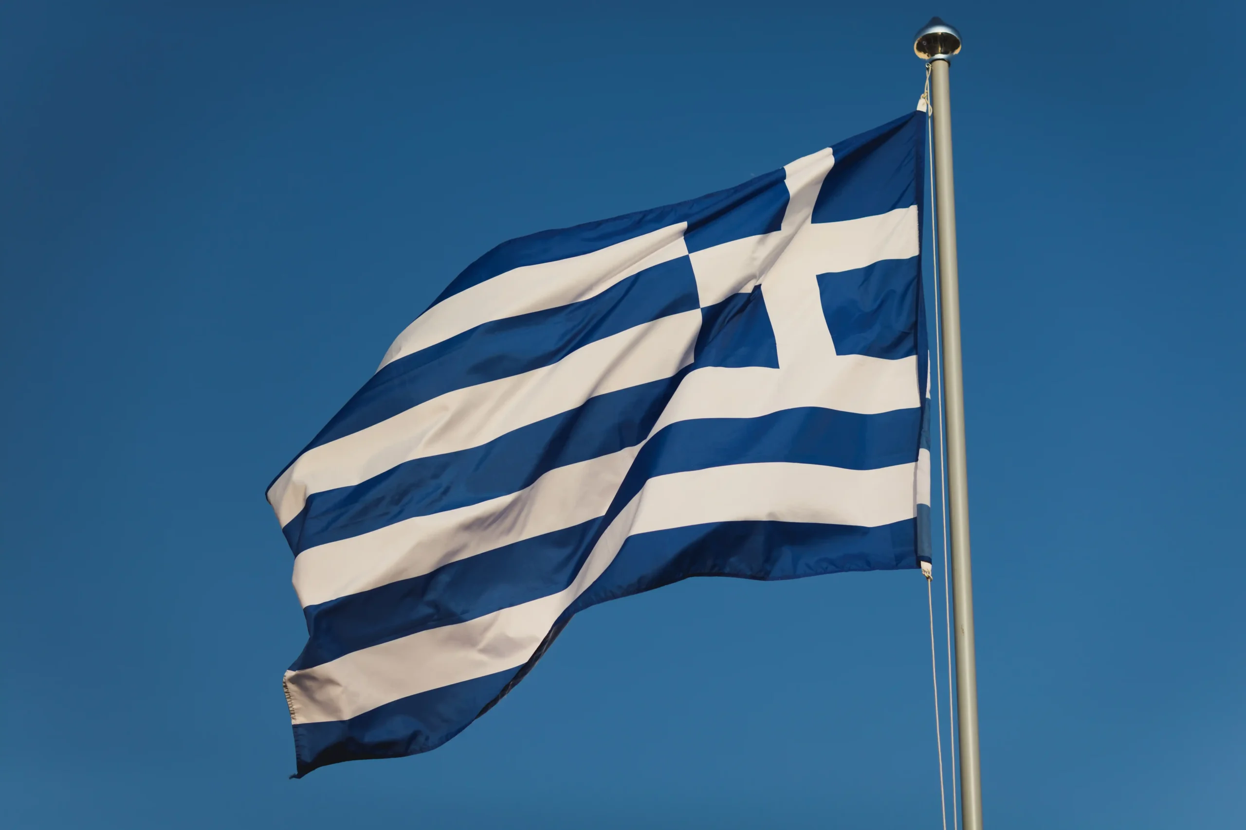 A imagem mostra a bandeira da Grécia hasteada, balançando com o vento. Ilustra o texto sobre Acordo Previdenciário Internacional Grécia.