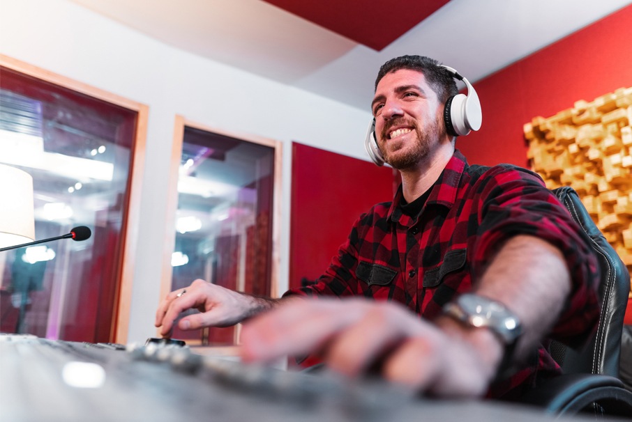 A imagem mostra um homem com fone, equalizando música no seu computador, em um estúdio de música. A imagem ilustra o texto 