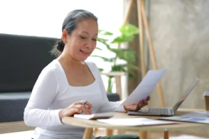 A imagem mostra uma mulher madura, sentada, em frente a uma escrivaninha, trabalhando com laptop, calculadora e documentos. Ilustra o texto sobre comprovante de profissional autônomo junto ao INSS.