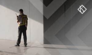A imagem mostra um vigilante parado de pé e de costas, usando um colete de trabalho, enquanto utiliza um rádio comunicador, que está perto de sua boca e ilustra a publicação 