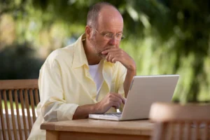 A imagem mostra um homem de meia-idade, sentado à mesa, utilizando um laptop.