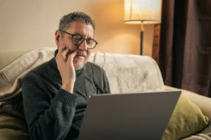 A imagem mostra um homem de meia-idade, sentado em seu sofá, dentro de casa, utlizando o laptop.