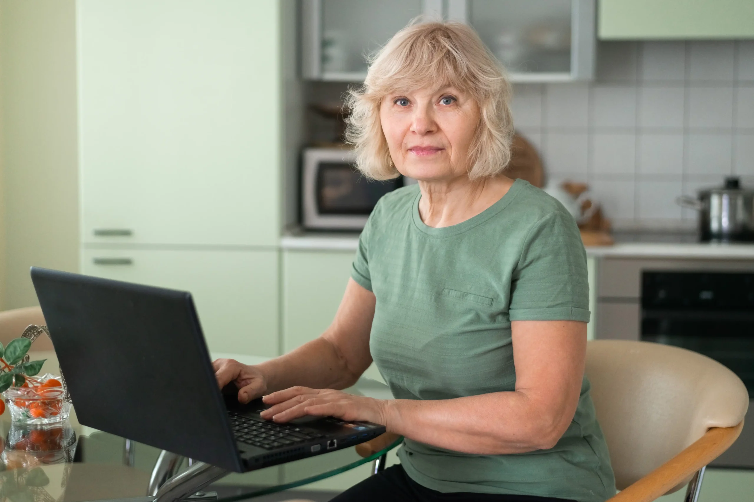 A imagem mostra uma sênior trabalhando no laptop sentada na cozinha, olhando para a câmera. Ilustra o texto sobre PPP, se a empresa fechou.