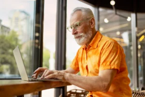 A imagem mostra um homem sênior, usando o laptop, sentando em uma cadeira, em um café. E ilustra o texto sobre Tempo fictício na aposentadoria de servidor público.