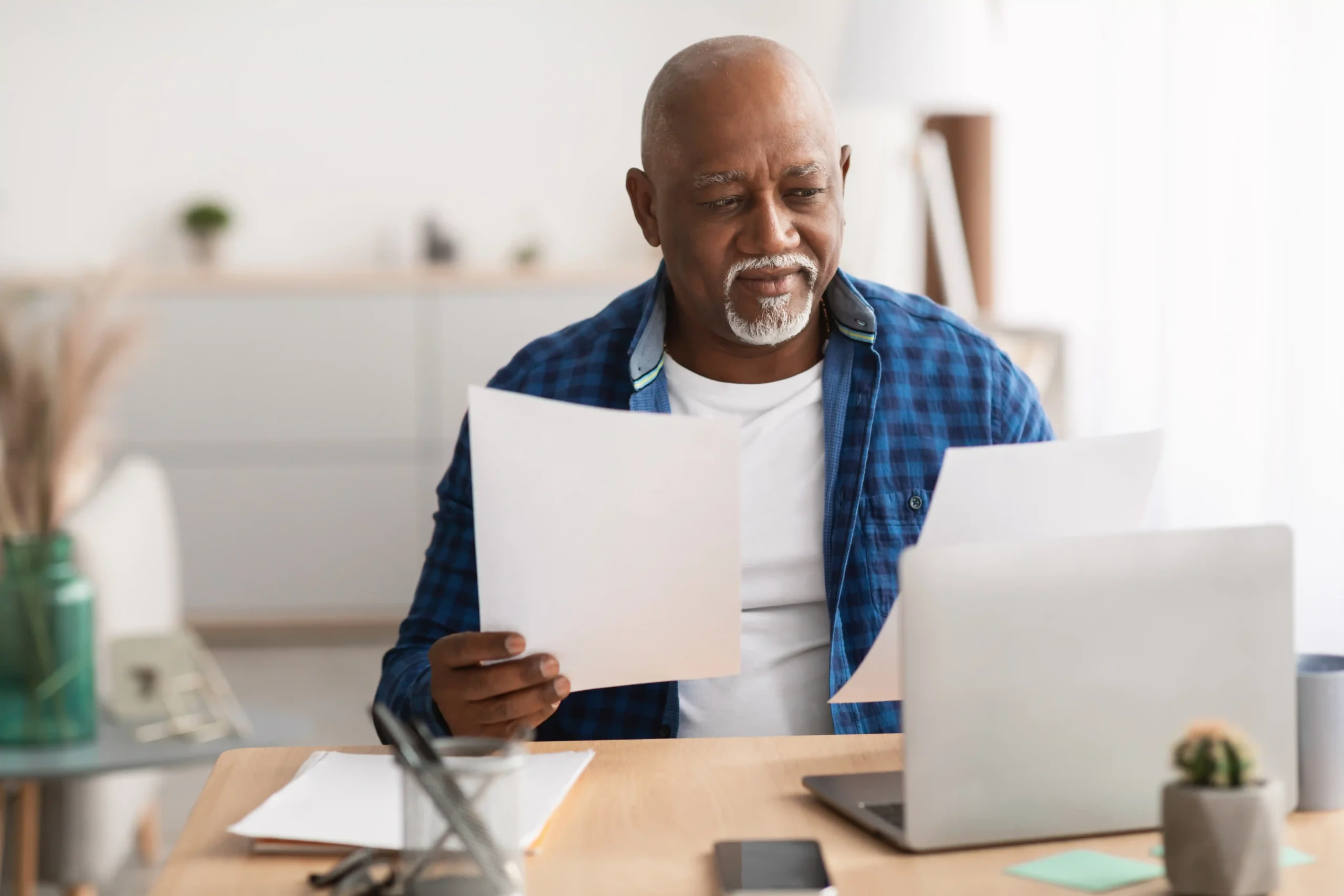 A imagem mostra um homem sênior, negro, usando o computador e segurando documentos com uma das mãos. Ilustrando o texto sobre como agendar perícia no INSS para funcionário.