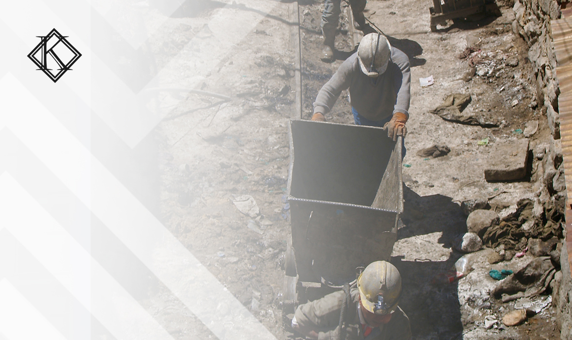 Dois homens empurram carrinho de mineração usando roupas protetoras, luvas e capacete em meio a uma mina. Sobre a imagem a identidade visual e logo da koetz advocacia. A imagem ilustra o texto 