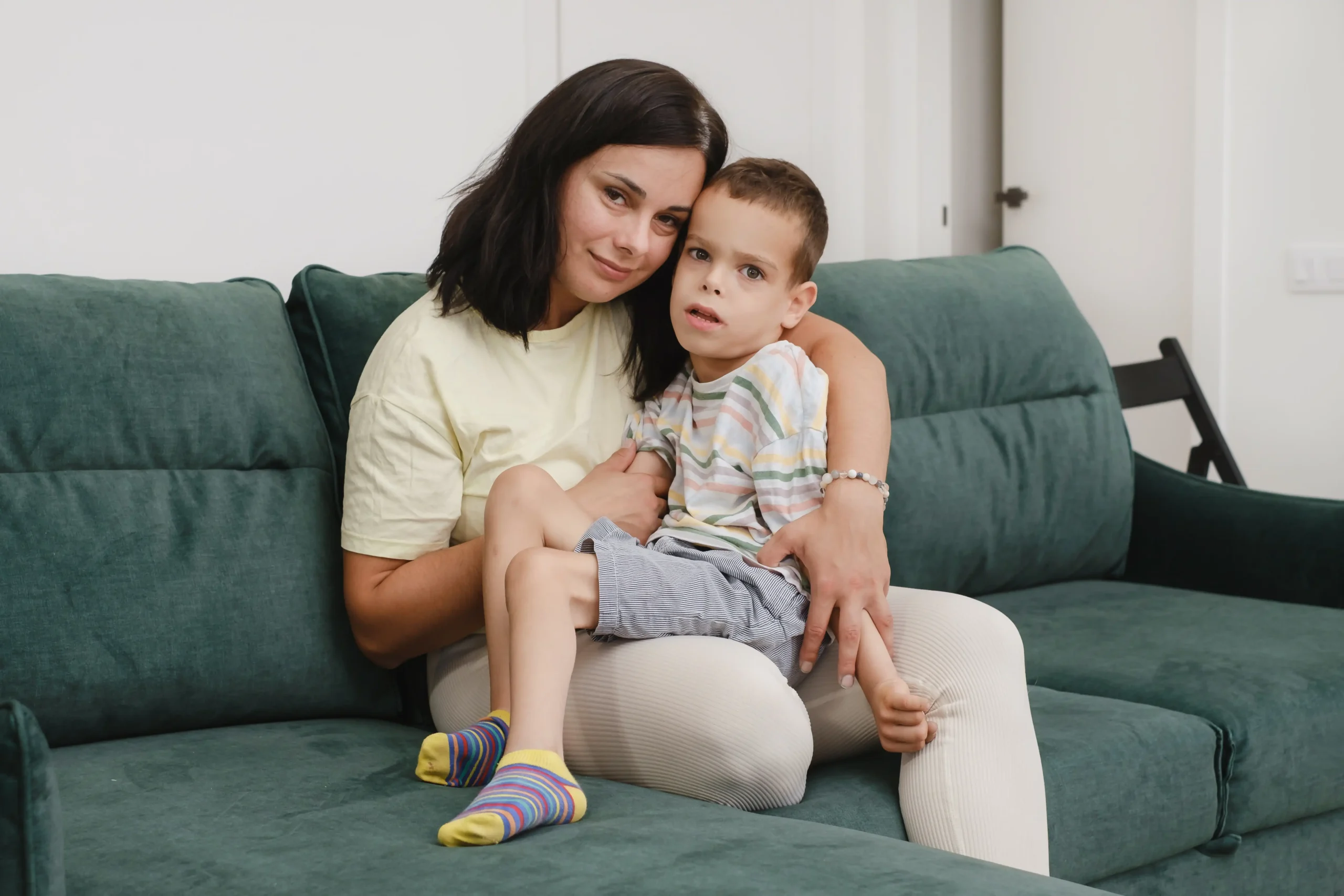 A imagem mostra uma mãe com filho com deficiência, com paralisia cerebral no sofá se divertindo. Ambos estão abraçados.