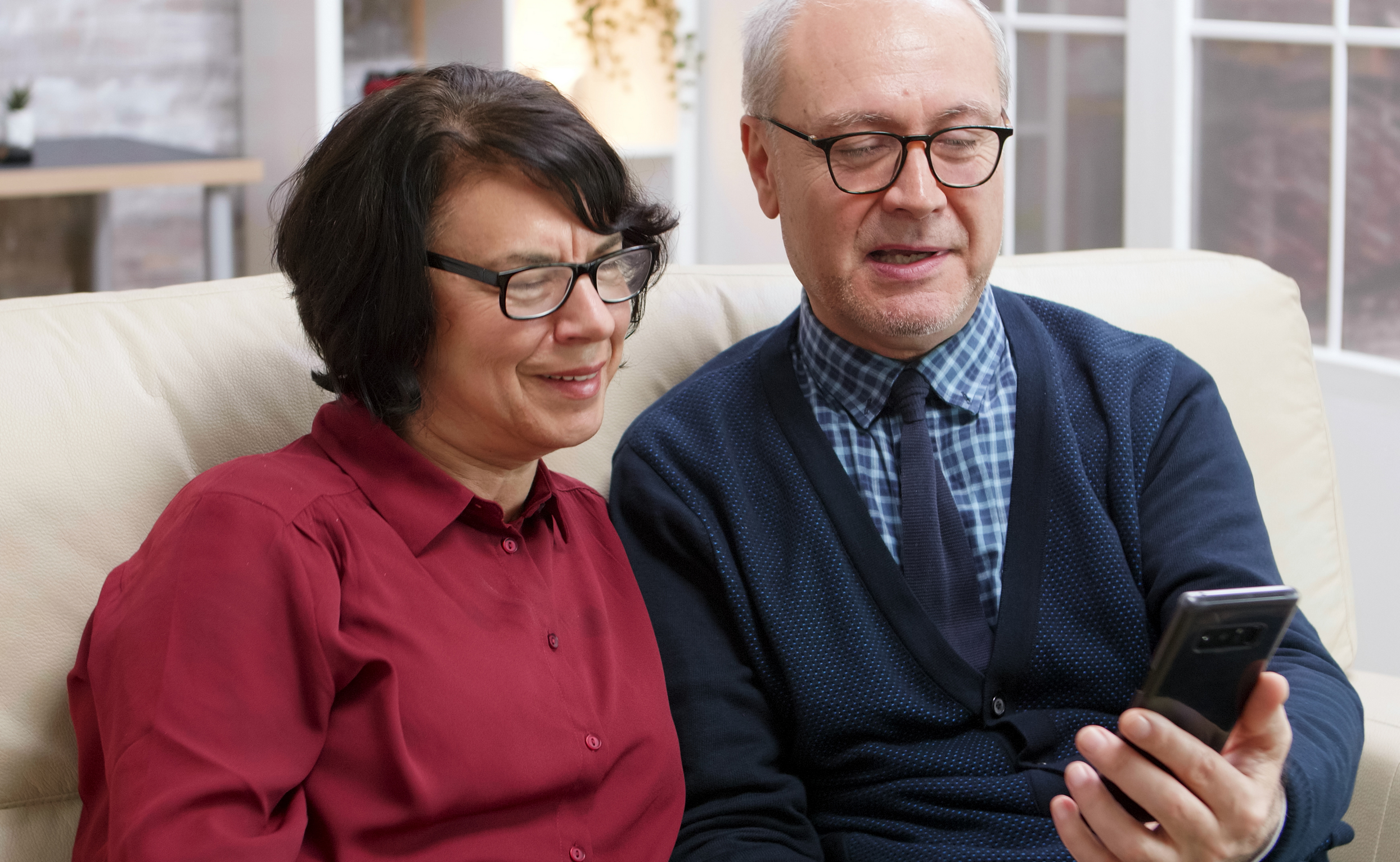 A imagem mostra um casal de idosos felizes, onde o homem está segurando um smartphone e ambos sorriem para e ilustra o texto: Aposentadoria no exterior: como contribuir, pedir e receber da Koetz Advocacia.
