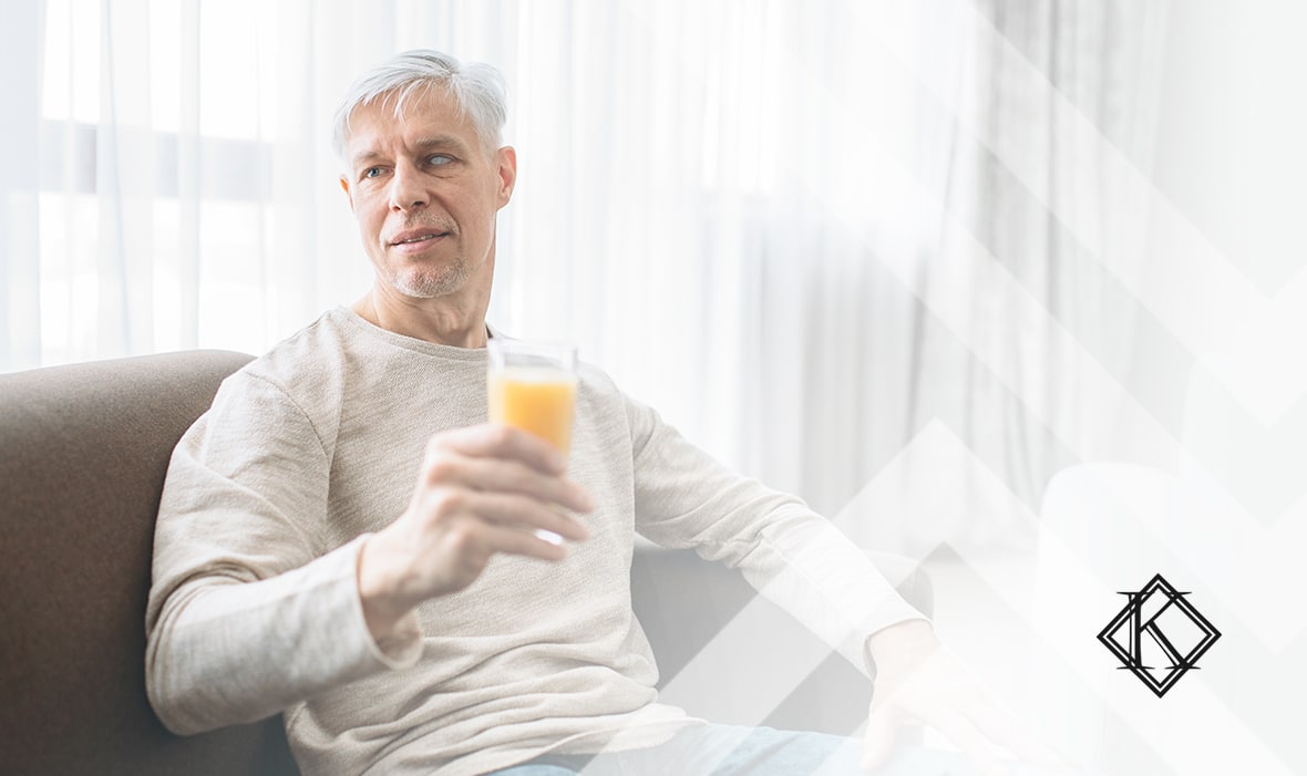 A imagem mostra um homem com visão monocular. Ele possui cabelos curtos e brancos, está sentado em um sofá, enquanto segura um copo cheio de suco de laranja e está olhando para o lado esquerdo, com um meio sorriso e ilustra a publicação 