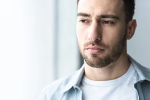 A imagem mostra um homem jovem, com barba, pensativo, olhando para longe em sua casa.