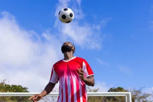 A imagem mostra um jogador de futebol negro, em uma camisa vermelha e branca equilibrando a bola de futebol na cabeça. E ilustra o texto sobre aposentadoria do jogador de futebol.