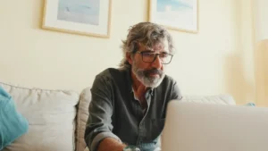 A imagem mostra um homem de meia-idade, sentado em seu sofá, dentro de casa e utilizando o laptop com uma expressão concentrada. Ilustra o texto sobre auxílio doença previdenciário.