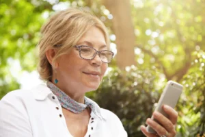 A imagem mostra uma mulher madura, ao livre, sorrindo, enquanto utiliza um smartphone. Ilustra o texto sobre consultar aposentadoria pelo CPF.