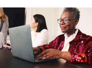 A imagem mostra uma mulher madura sorrindo, com seu computador. A imagem ilustra o texto 