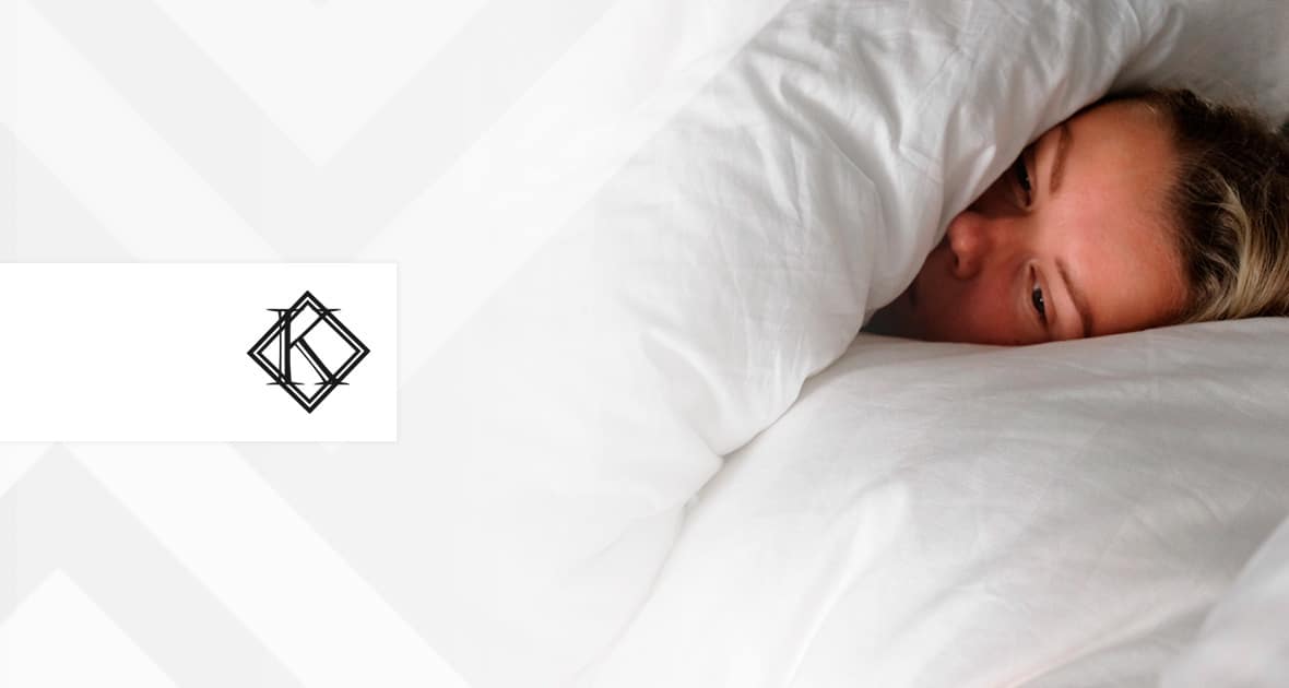 A imagem mostra uma mulher doente na cama, com expressão séria. Ela está coberta por um edredom, enquanto deitada em uma cama com lençóis e travesseiros brancos, e ilustra a publicação 