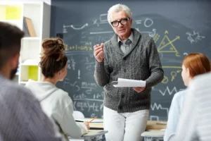 A imagem mostra um professor sênior, de cabelos curtos e brancos, em sala de aula, com seus alunos. Ilustra o texto sobre valor da aposentadoria do professor.