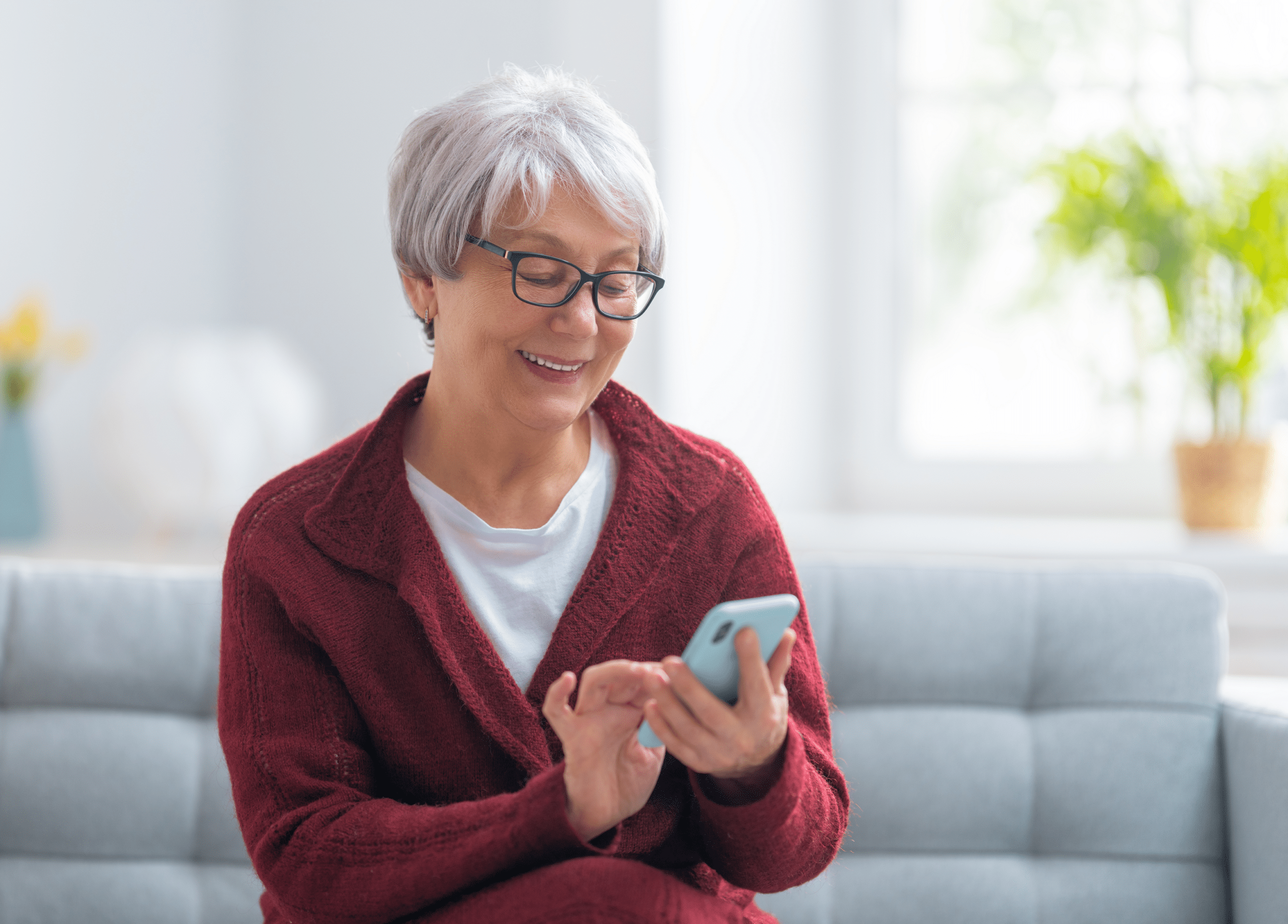 A imagem mostra uma mulher idosa, sentada em um sofá, sorrindo, de óculos, enquanto segura um smartphone com uma das mãos e mexe na tela com a outra. Além disso, ela está sentada em um sofá, dentro de casa e ilustra o texto: Averbação no INSS: como fazer, prazos, regras e opções! da Koetz Advocacia.