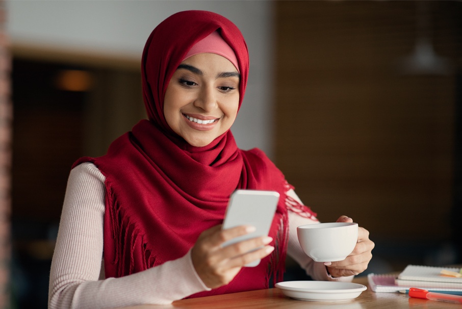 A imagem mostra uma mulher sorrindo enquanto olha para a tela do seu celular, e a outra mão está segurando uma xicara. Imagem do texto