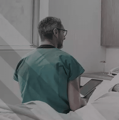 A imagem mostra um enfermeiro, com roupas hospitalares, segurando um tablet. Ele está de costas, sentado em uma cama hospitalar, perto de um paciente.