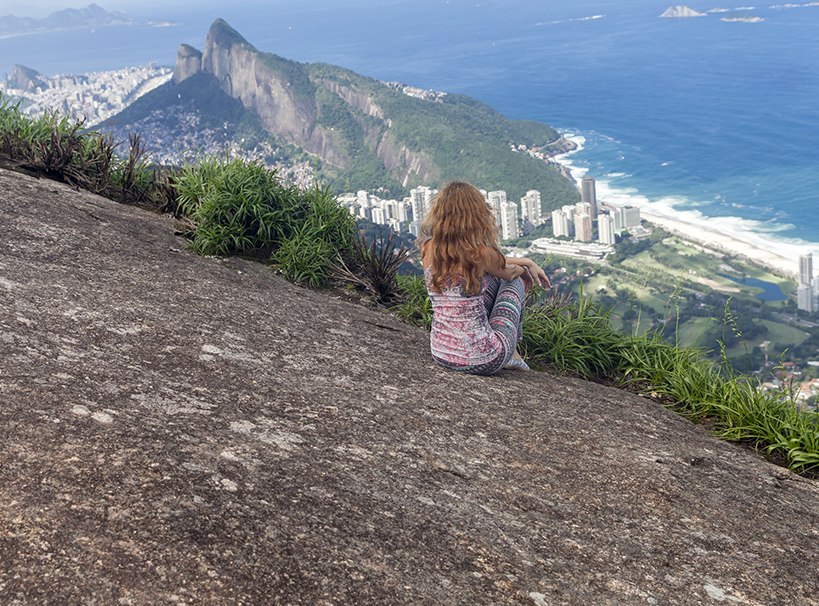 A imagem mostra uma mulher, sentada de costas para a câmera, olhando de frente para a vista do Rio de Janeiro, enquanto está sentada em uma pedra.