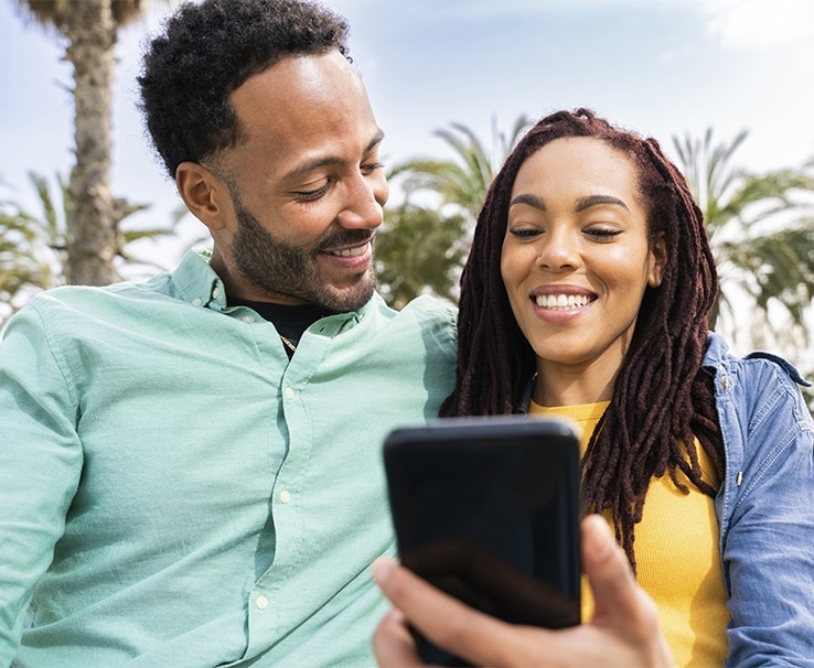 A imagem mostra um jovem casal negro, lado a lado, olhando para o celular, que está nas mãos da mulher.