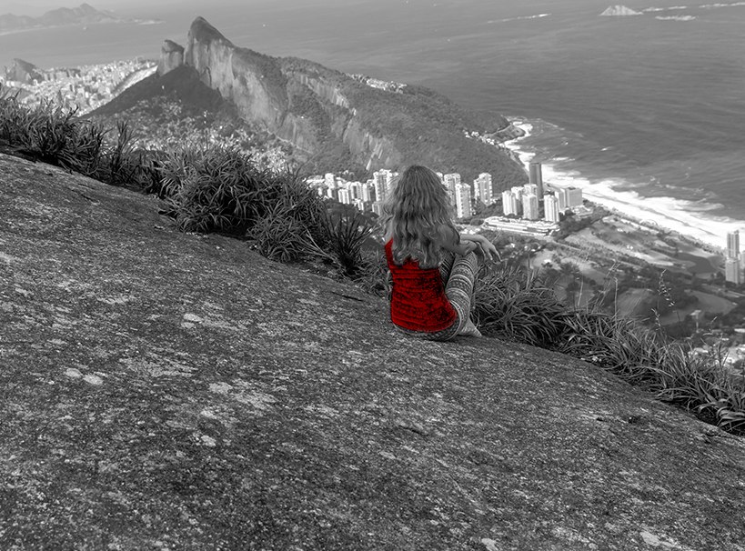 A imagem mostra uma mulher, sentada de costas para a câmera, olhando de frente para a vista do Rio de Janeiro, enquanto está sentada em uma pedra.