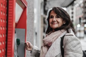 A imagem mostra uma mulher adulta pegando dinheiro de uma caixa eletrônico na rua. A imagem ilustra o texto 