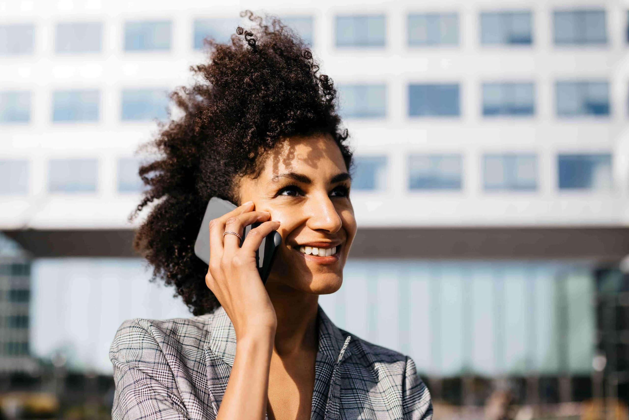 A imagem mostra uma mulher, falando ao celular, enquanto sorri. Ela está ao ar livre.