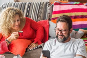 A imagem mostra uma mulher em um sofá com uma xícara de café, perto de um homem olhando o celular e ambos sorriem e ilustra o texto: Types of residence permits in Brazil with a fixed term da Koetz Advocacia.