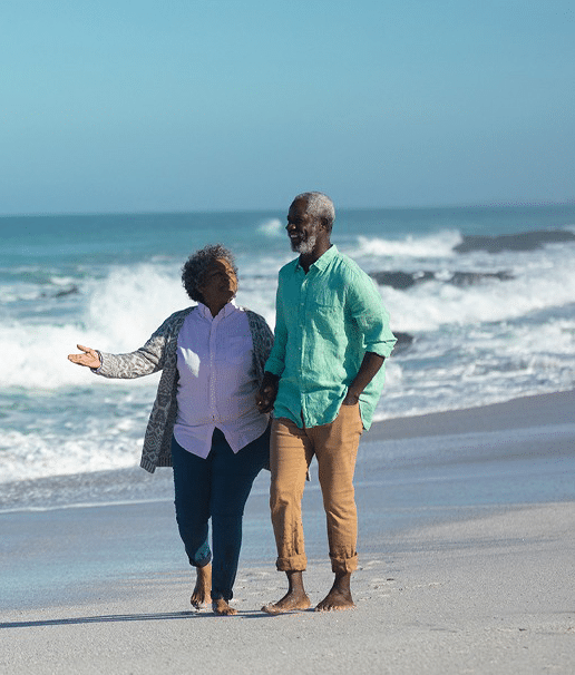 A imagem mostra um casal de idosos negros, caminhando na praia, de mãos dadas.