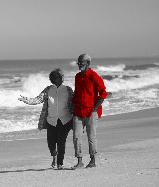 A imagem mostra um casal de idosos negros, caminhando na praia, de mãos dadas.