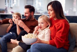 A imagem mostra um casal na sala, com seus filhos no colo, enquanto comem pipoca. A imagem ilustra o texto 
