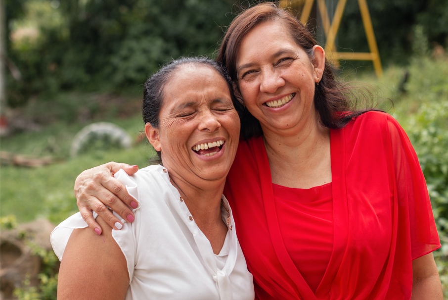 La imagen muestra a dos mujeres adultas riéndose y abrazándose. La imagen ilustra el texto "Naturalización brasileña con 1 año de residencia permanente" de Koetz Advocacia