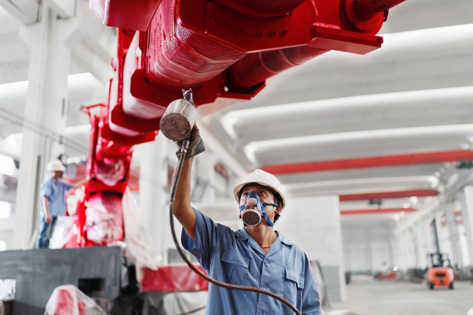 A imagem mostra um homem chinês pulverizando a pintura de um braço de guindaste vermelho na oficina de uma fábrica e ilustra o texto: Comércio Brasil e China da Koetz Advocacia.