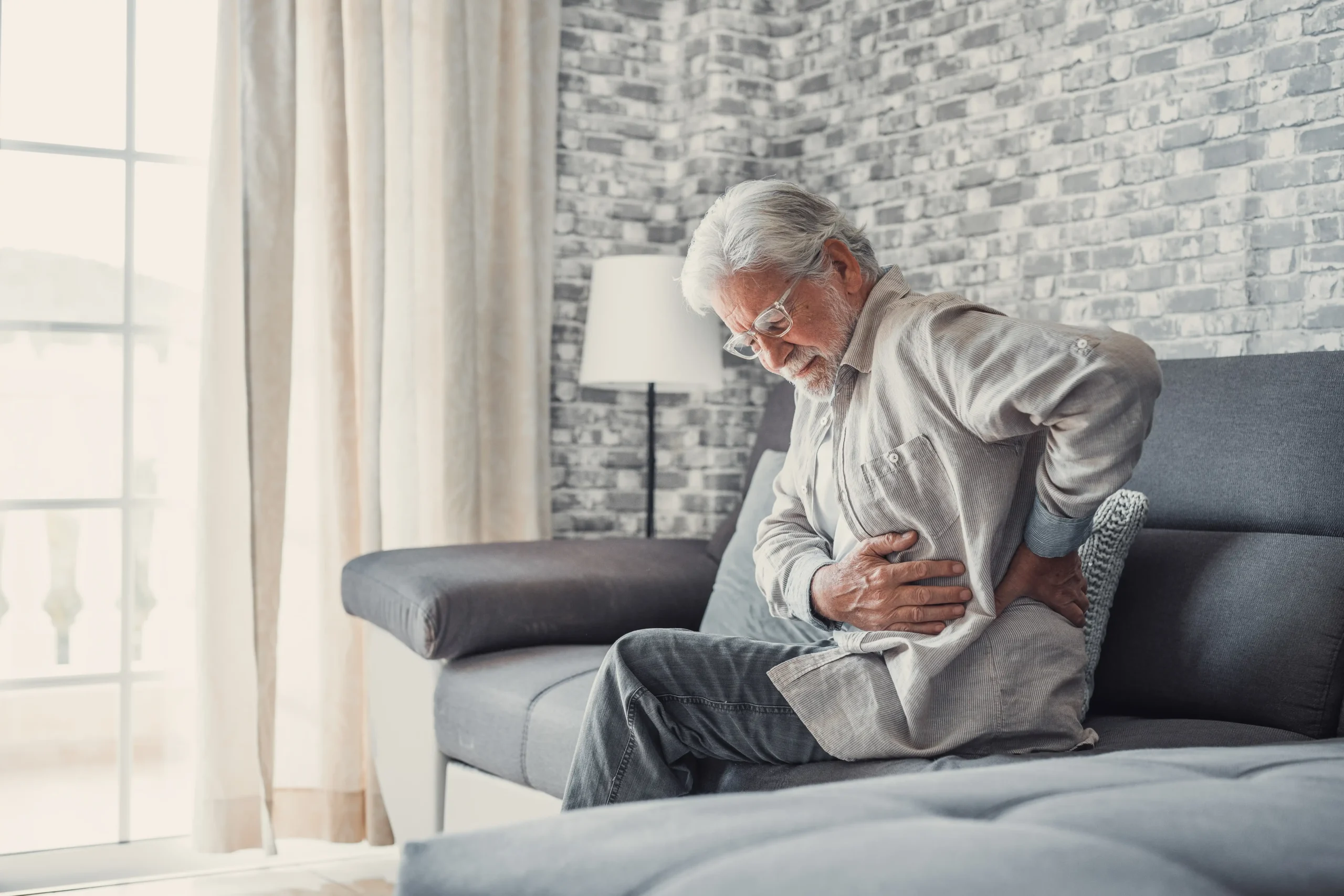 A imagem mostra um homem sênior, sentado na cama, com dores nas costas. E ilustra o texto sobre Nexo causal e concausal.