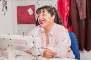 La imagen muestra a una mujer madura sonriente sentada a una mesa con una máquina de coser. ilustrando el articulo 