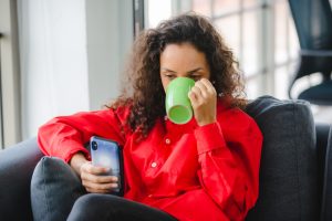 A imagem mostra uma jovem mulher sentada em um sofá, bebendo café em uma xícara e olhando um smartphone e ilustra o texto: Volta da exigência de visto de turista para americanos da Koetz Advocacia.