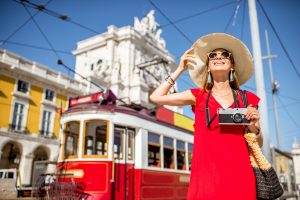 A imagem mostra uma jovem mulher andando por Lisboa, Portgual, enquanto sorri e segura uma câmera fotográfica e ilustra o texto: Tipos de Vistos para Portugal da Koetz Advocacia.